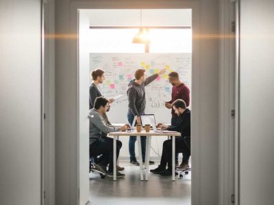 Team collaborating on a project inside an office viewed through a slightly open hallway door with sticky notes and whiteboard brainstorming.